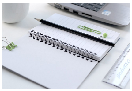 Em cima de uma mesa há um caderno em branco, um notebook, uma régua e uma caneca