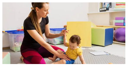 The image shows a physiotherapist helping a baby to sit up, both are on the floor